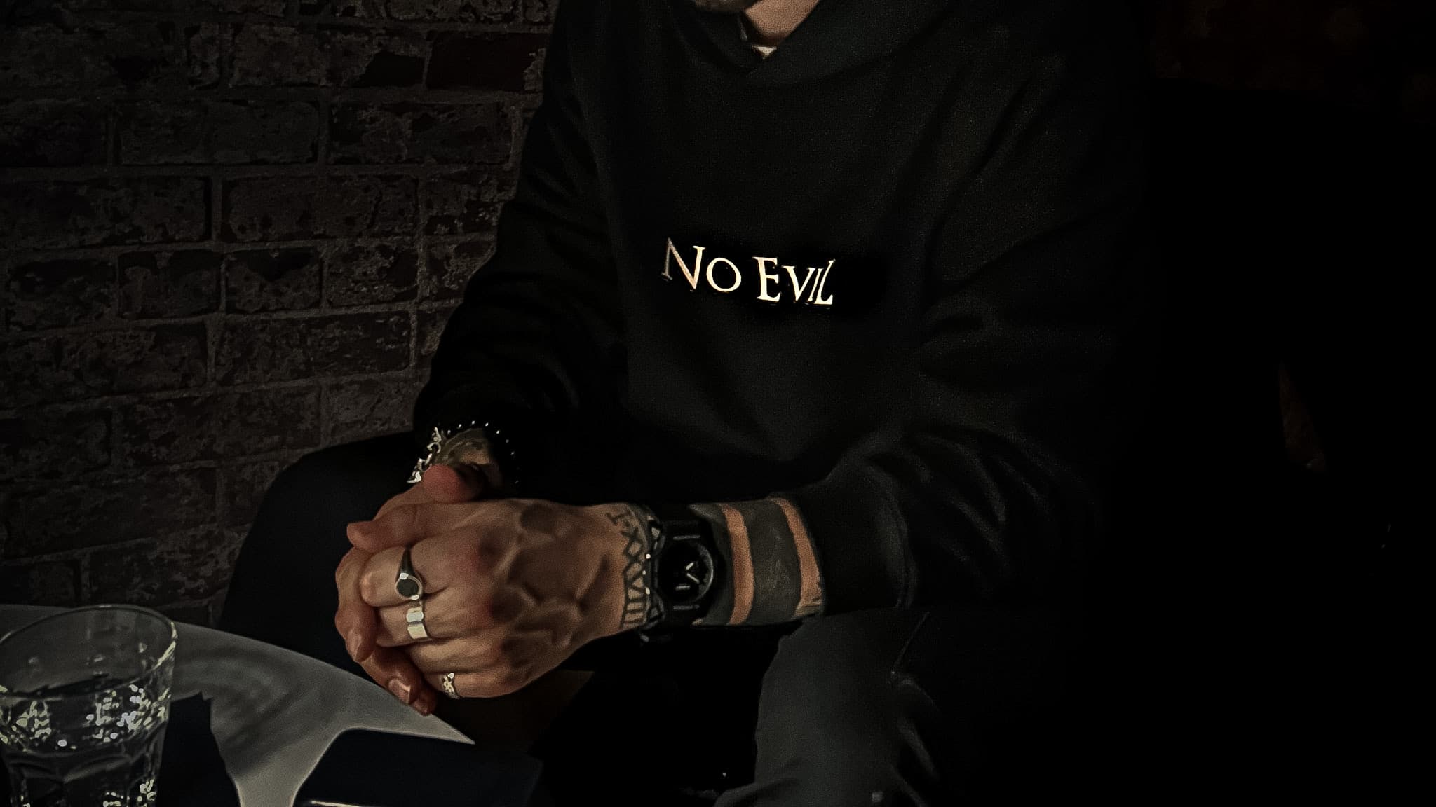 Model in a No EviL hoodie with clasped hands at a table in low light